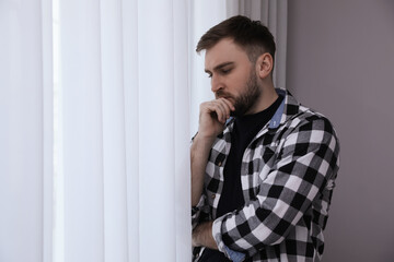 Young depressed man near window at home, space for text