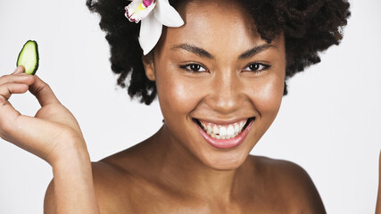 Smiling african american woman with orchid in hair holding slice of cucumber isolated on grey