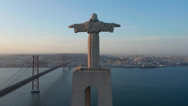 Drone camera ascending along pedestal and statue. Evening back view of Christ the King Sanctuary in Almada. Long cable-stayed bridge over Tagus river in background. Lisbon, capital of Portugal.