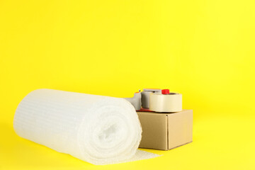 Bubble wrap roll, tape dispenser and cardboard box on yellow background, space for text