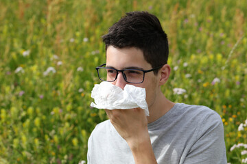 myopic boy with glasses sneeze because of allergies to grass on flowery field in Spring