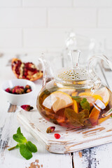 Fruit tea with berries, lemon, lime and mint leaves in glass teapot on white light wooden background. Selective focus
