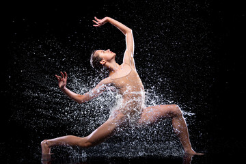 Wet young woman in tight beige swimsuit is dancing on floor under rain and splashes, drops of...