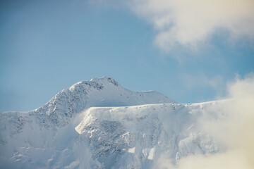 Minimalist view of snow-capped mountain wall in thick low clouds in sunshine. Scenic bright mountain landscape with white-snow peak among dense clouds in blue sky. Wonderful scenery with snowy top.