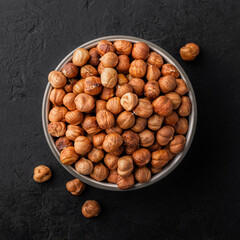 Peeled hazelnuts in a bowl and a few nuts scattered around on a black textured background, top view.