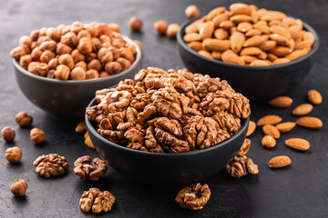 Different types of peeled nuts in bowls and a few nuts scattered around on a black textured background, side view.