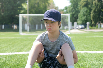 Boy sitting soccer field in summer day. Leisure children with Down syndrome, disability