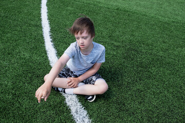 Young boy sitting soccer green field in summer day. Сhildhood сhildren with Down syndrome, disability