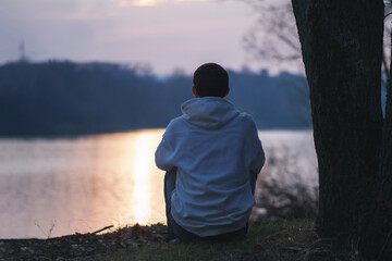 alone young man in a white hoodie sits on the riverbank by a tree at sunset