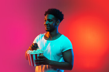 Image of young african man with the box of popcorn in his hands on bright pink neon background.