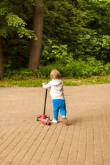 A preschool child rushes on a scooter through the park. Baby in the park in sunny weather. The boy has fun walking in the park
