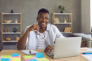 Portrait of young smiling cheerful african american businessman startup project manager entrepreneur with eyeglasses sitting at office home desk front of laptop looking directly at camera