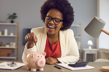 Happy black woman puts coin in pink piggy bank. Smiling cheerful businesswoman sitting at office...
