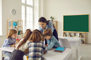 Young caucasian female teacher helping children with their task in classroom at elementary school. Group of focused children sit together at a desk and watch and listen carefully to the teacher.