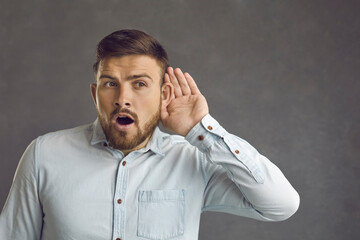 Omg I can hear them talk about something interesting. Curious attentive guy isolated on gray background puts hand to ear overhearing top secret information or listening to hot news and shocking gossip