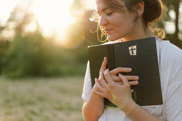 Christian woman holds bible in her hands. Reading the Holy Bible in a field during beautiful sunset. Concept for faith, spirituality and religion