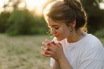 Woman closed her eyes, praying in a field during beautiful sunset. Hands folded in prayer concept for faith, spirituality and religion. - obrazy, fototapety, plakaty