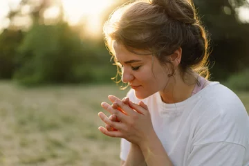 Fotobehang Woman closed her eyes, praying in a field during beautiful sunset. Hands folded in prayer concept for faith, spirituality and religion. © Анастасія Стягайло