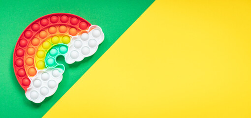 Multicolored silicone toy antistress pop it in the shape of a rainbow with clouds on a green yellow diagonal background - obrazy, fototapety, plakaty