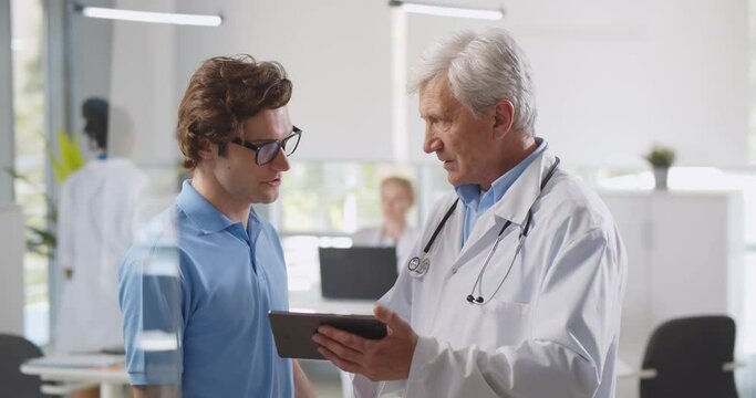 Mature male doctor pointing at digital tablet screen sharing health tests results to patient