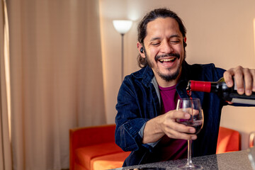 Young latin man serving red wine on a glass at home in the living room
