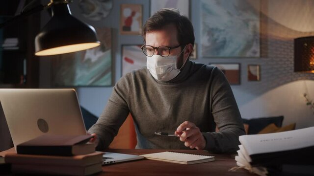 Experienced man doctor wearing medical mask and glasses, professional psychologist, coach typing on laptop computer, writing notes. Remote office during coronavirus covid 19 lockdown.