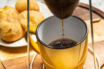 Café sendo coado no coador com uma caneca amarela abaixo com um prato de pão de queijo ao fundo.