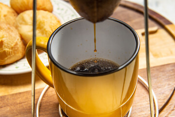 Café sendo coado no coador com uma caneca amarela abaixo com um prato de pão de queijo ao fundo.