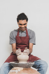 smiling young african american man in casual clothes and apron modeling wet clay pot on wheel in pottery