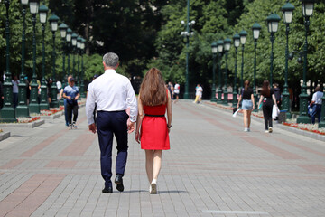 Gray-haired man in business clothes walking with young slim girl in red dress on the street in summer city