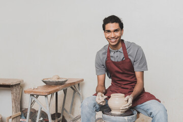 smiling young african american man shaping wet clay pot on wheel with scraper in pottery