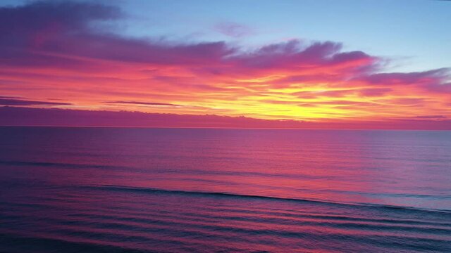 Aerial of dramatic predawn sky with colors of red, orange, yellow, and purple over a magenta and dark purple ocean.