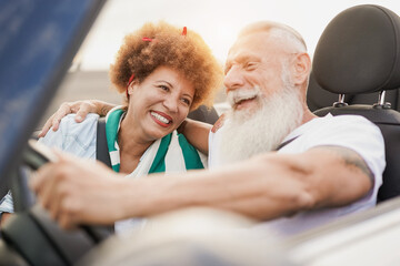 Trendy mature couple having fun inside convertible car - Multiracial senior people on a road trip...
