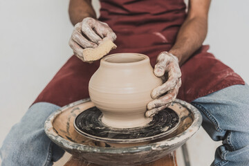 partial view of young african american man sitting on bench and shaping wet clay pot on wheel with sponge in pottery