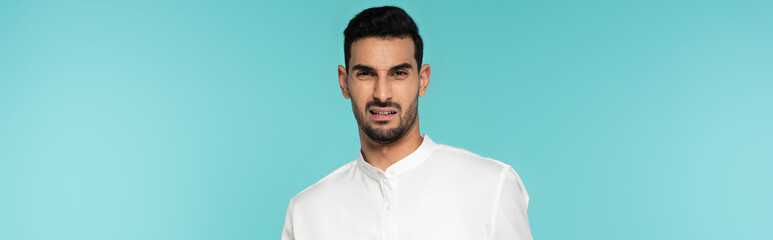 Confused arabian man in white shirt looking at camera isolated on blue, banner