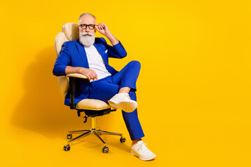 Portrait of attractive imposing serious man sitting in chair touching specs isolated over bright yellow color background