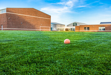 Old and deflate orange football on green turf with sunlight at school building background, depth of field. Outdoor recreation space for students. Landscape view of empty soccer field and unused ball.