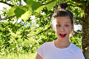 in summer garden cute cheerful girl in white T-shirt holds ripe cherry berry in her mouth. Delicious and healthy food. Close-up