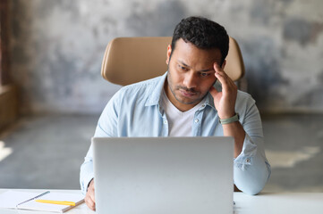 Exhausted indian office employee sits at the desk with a laptop and holds head with hand, tired guy...