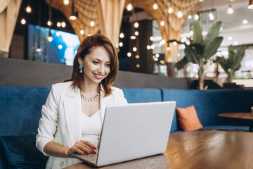 Beautiful Young Freelancer Woman Using Laptop Computer Sitting At Cafe Table. Happy Smiling Girl Working Online Or Studying And Learning While Using Notebook. Freelance Work, Business People Concept