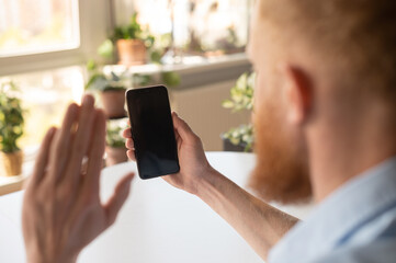 Video call concept. A man is holding mobile phone and waving hi, making video call, phone with blank empty screen, using new mobile app for video connection, back view, copy space, template