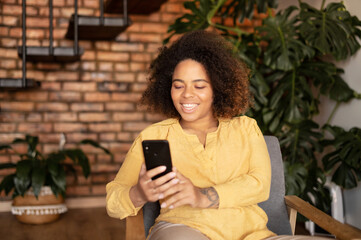 Joyful young African-American woman using smartphone sitting on the armchair at home, smiling black female enjoys chatting online, spending time in social networks