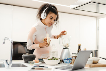 Obraz na płótnie Canvas Black woman in headphones using laptop while making smoothie at home