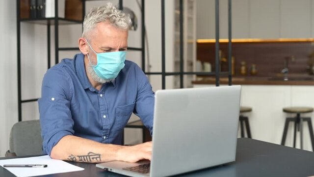 New reality and new rules for office staff during pandemic. Mature gray-haired male employee wearing medical mask is using laptop at the office, pandemic prevention and virus outspread