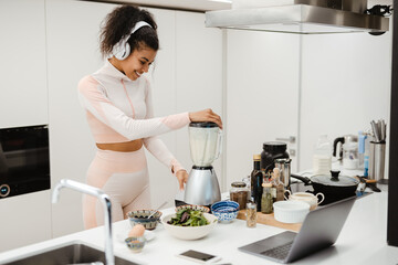 Black woman in headphones using laptop while making smoothie at home