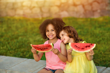 Funny Black girl and European girl are eating watermelon outdoors in the hot summer. Smiling children, healthy food