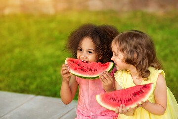 Funny Black and European girls are eating watermelon outdoors in the hot summer. Smiling children enjoy healthy food - Powered by Adobe
