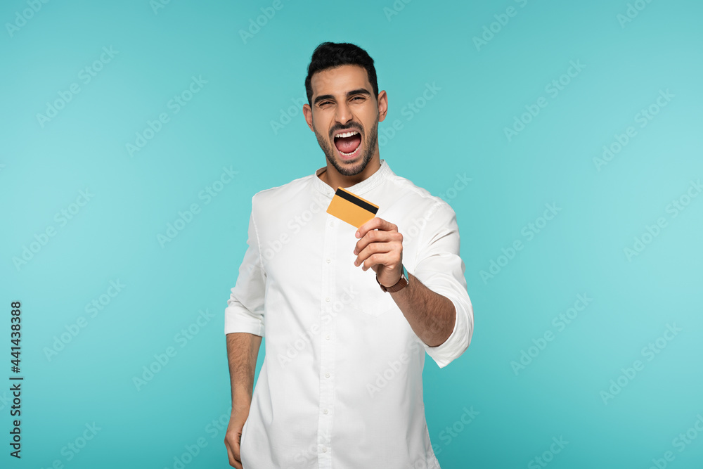 Wall mural excited arabian man showing credit card isolated on blue