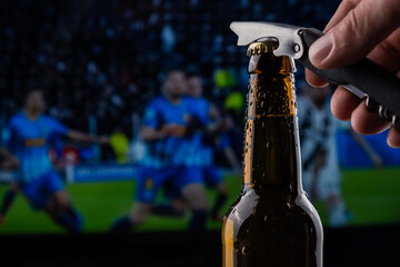 A man opens a bottle of beer in the background of a broadcast of a football match.