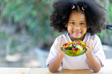 Little African American curly haired girl sits and eats vegetable. delicious salad on the table Fun, cheerful. Appetite. Childhood and eating concepts.